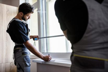 Two Professional blind installation Workers In Uniform Using Tape Measure while installing blinds